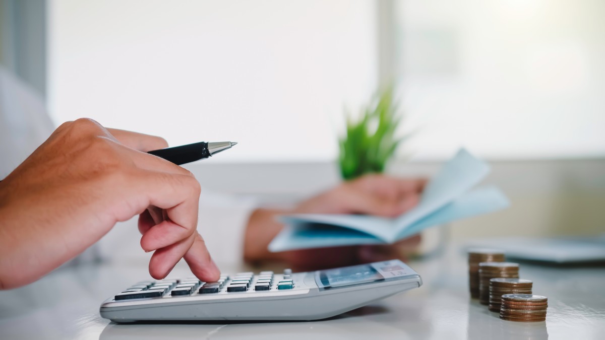 Close-up of a person using a calculator with coins stacked on a desk and holding a checkbook in the background.