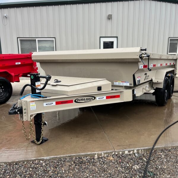 Beige Dura-Haul dump trailer with tandem black wheels and reflective tap along the frame, parked on wet concrete in front of a metal building with green trim