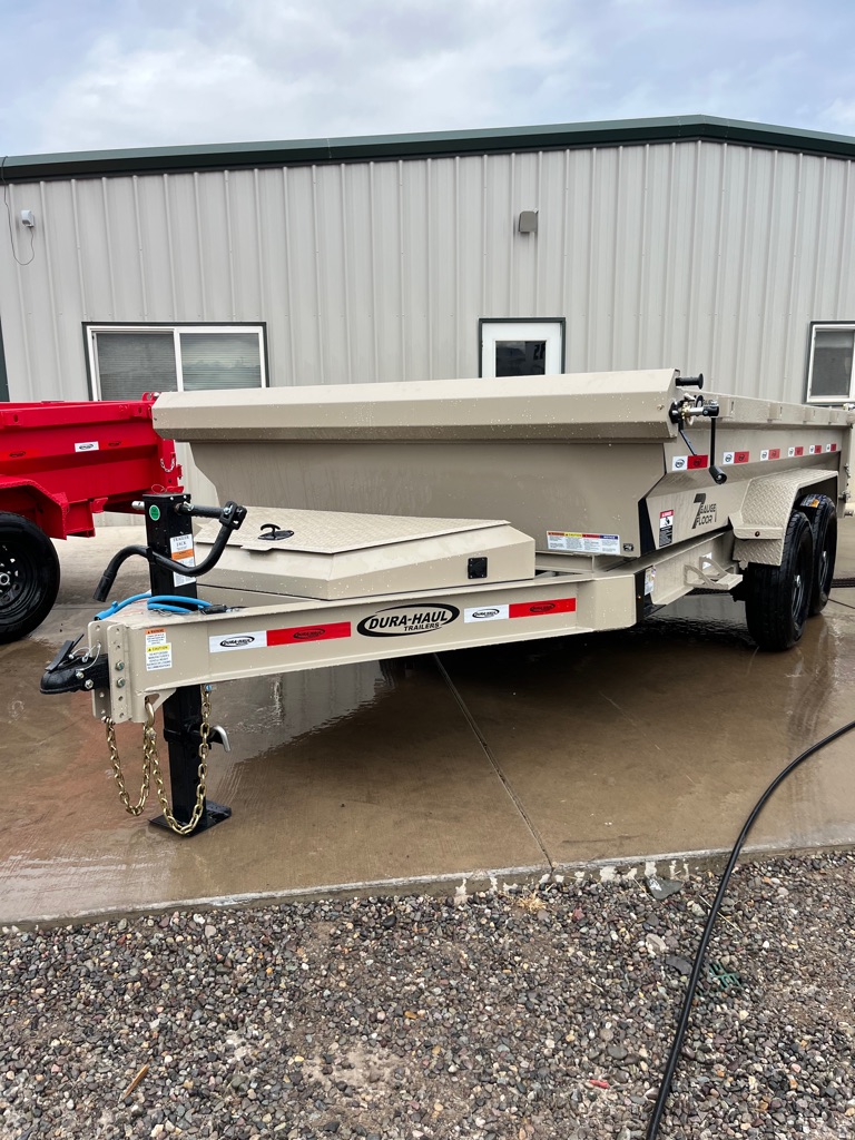 Beige Dura-Haul dump trailer with tandem black wheels and reflective tap along the frame, parked on wet concrete in front of a metal building with green trim
