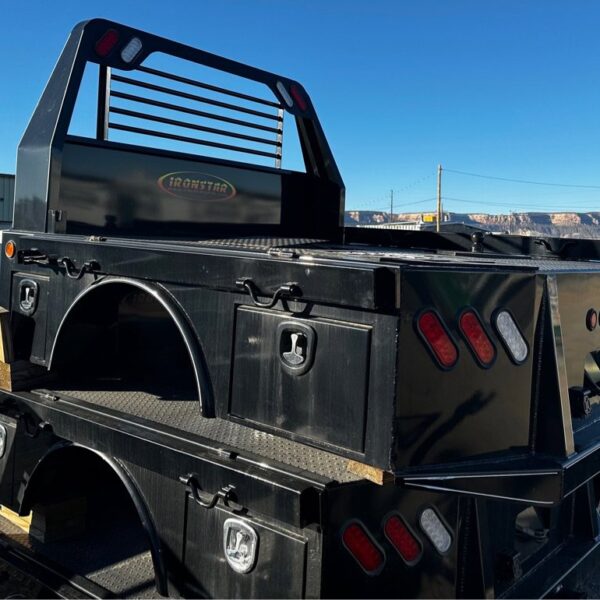 A black Ironstar flatbed truck bed with integrated toolboxes, red and white tail lights, and a protective headache rack, stacked on similar beds in an outdoor lot