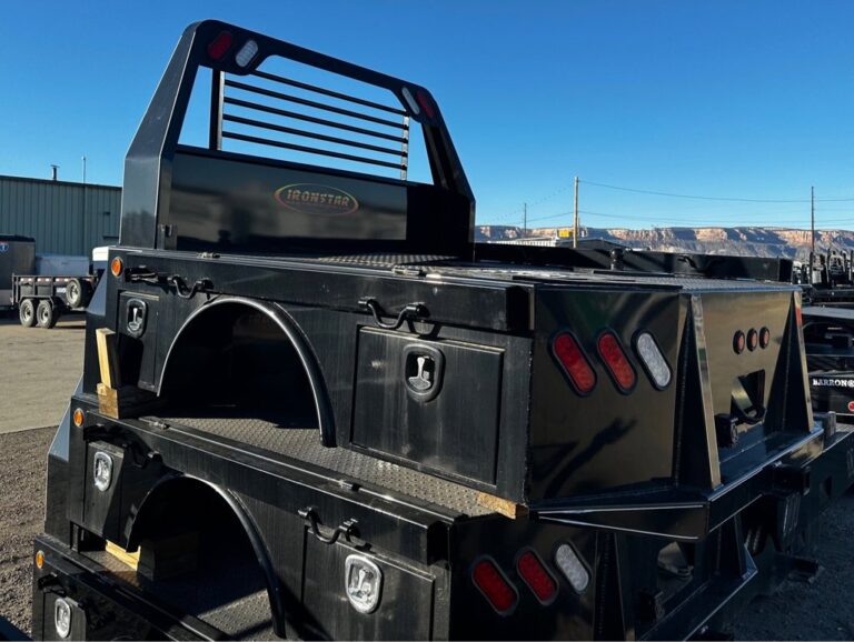 A black Ironstar flatbed truck bed with integrated toolboxes, red and white tail lights, and a protective headache rack, stacked on similar beds in an outdoor lot
