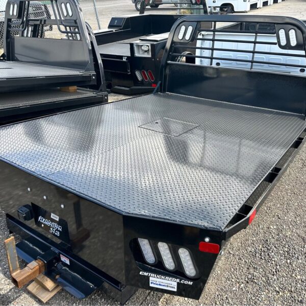 Two black CM flatbed truck beds with diamond plate surfaces and headache racks, parked on gravel lot