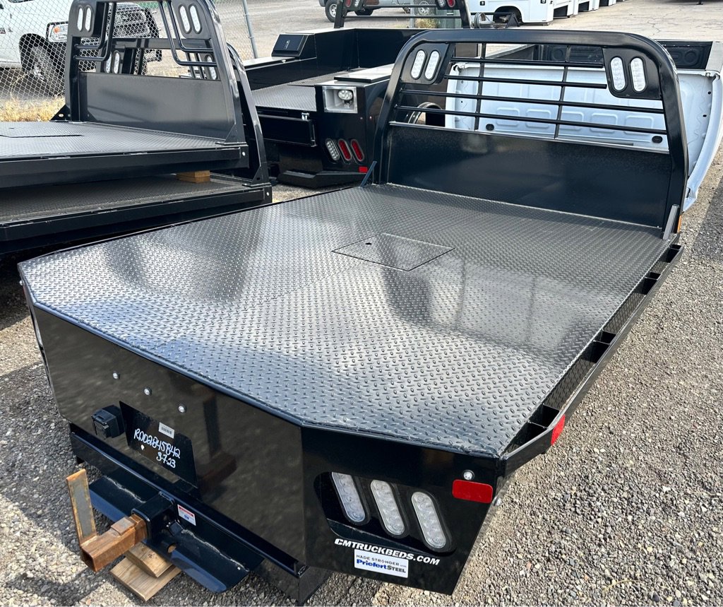 Two black CM flatbed truck beds with diamond plate surfaces and headache racks, parked on gravel lot