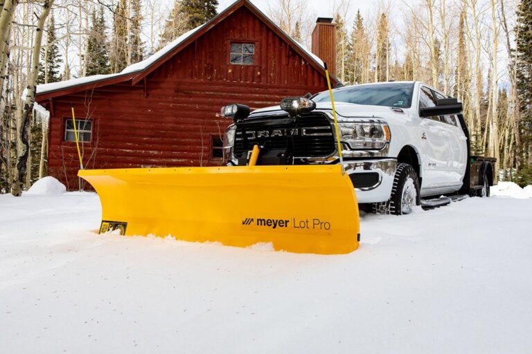 White truck with yellow Meyer Lot Pro snow plow clearing snow in front of red log cabin.