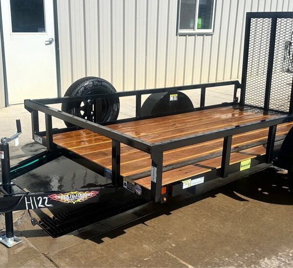 Small black utility trailer with wooden deck, rear mesh ramp, and single axle, parked on wet concrete near an office building