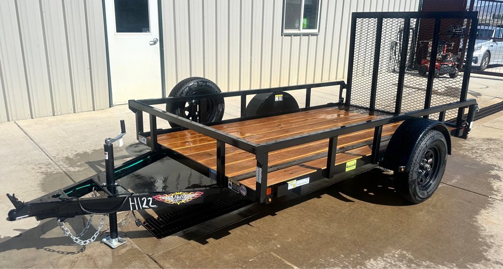 Small black utility trailer with wooden deck, rear mesh ramp, and single axle, parked on wet concrete near an office building