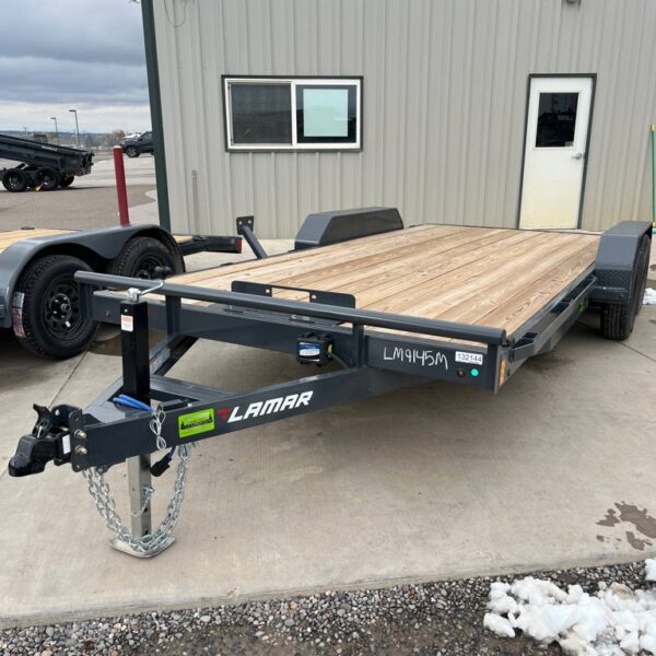 Black Lamar flatbed trailer with wood deck and tandem axles parked on concrete near metal office building
