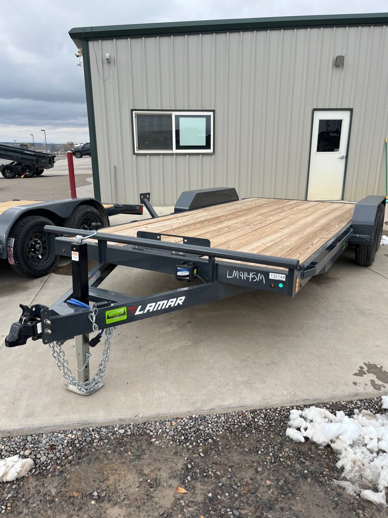 Black Lamar flatbed trailer with wood deck and tandem axles parked on concrete near metal office building