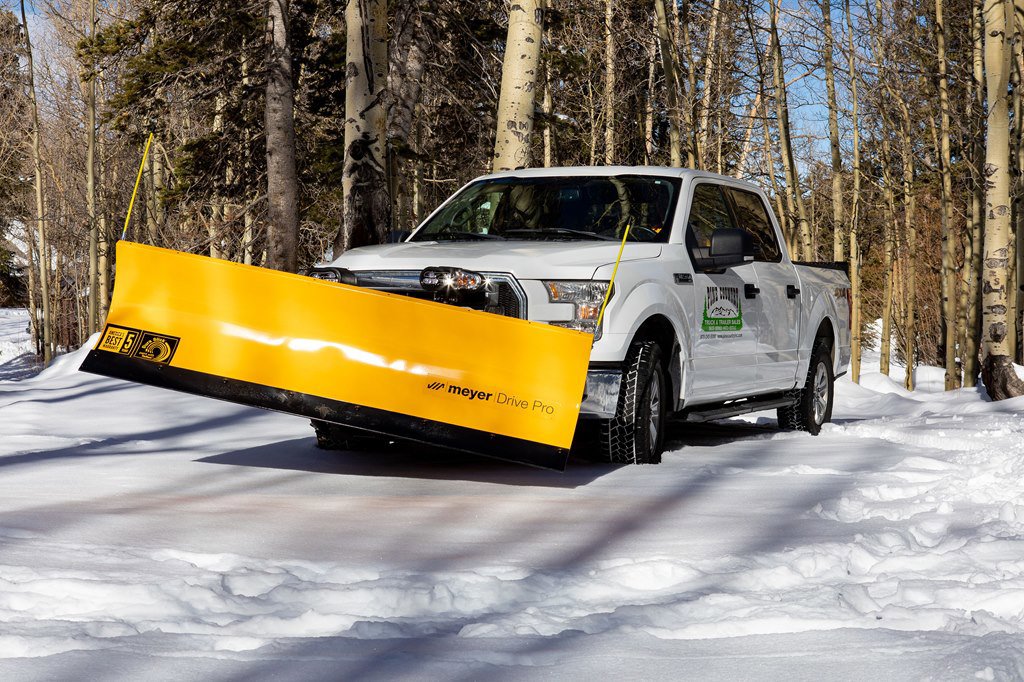 White pickup truck with yellow Meyer Drive Pro snow plow driving on snowy forest road lined with tall leafless trees.