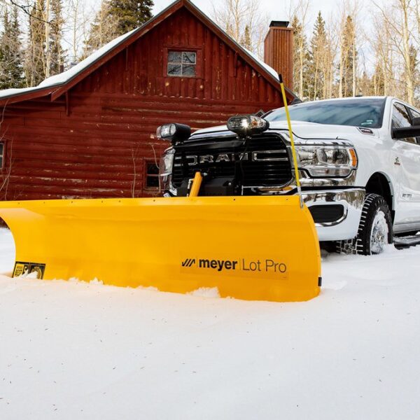 White truck with yellow Meyer Lot Pro snow plow clearing snow in front of red log cabin.