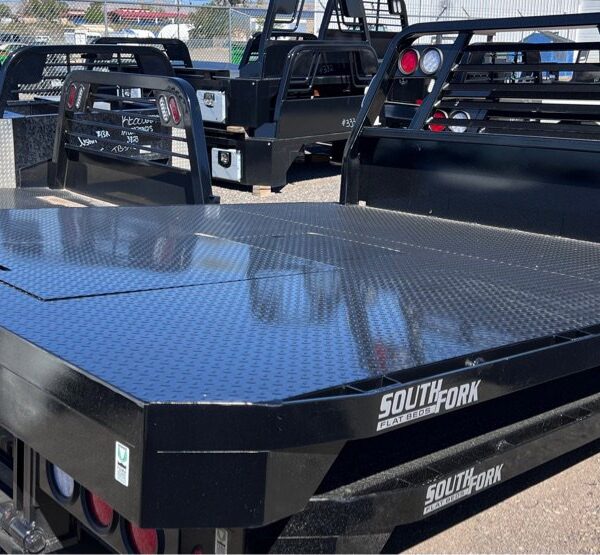 A black South Fork flatbed truck bed with a diamond-plate steel surface and integrated headache rack, displayed outdoors among other truck beds in a lot