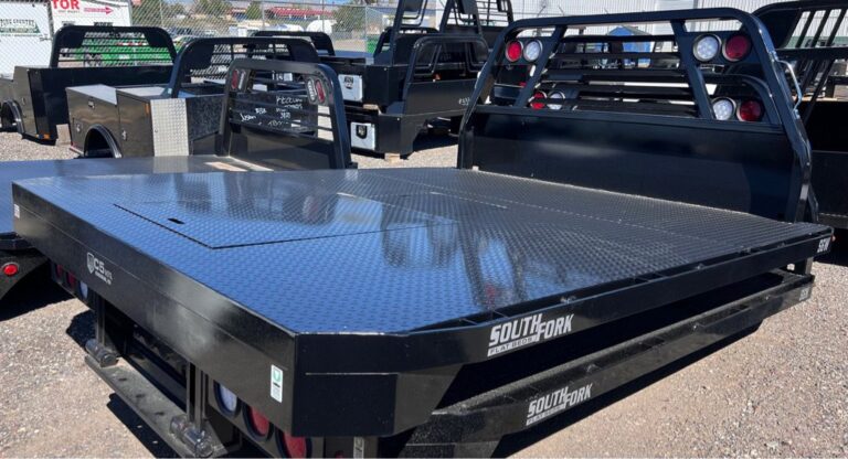 A black South Fork flatbed truck bed with a diamond-plate steel surface and integrated headache rack, displayed outdoors among other truck beds in a lot