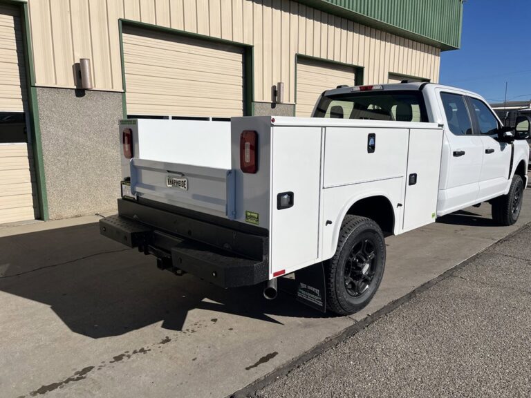 A white pickup truck with a utility service body, black wheels, and multiple storage compartments, parked outside a building with tan and green metal siding