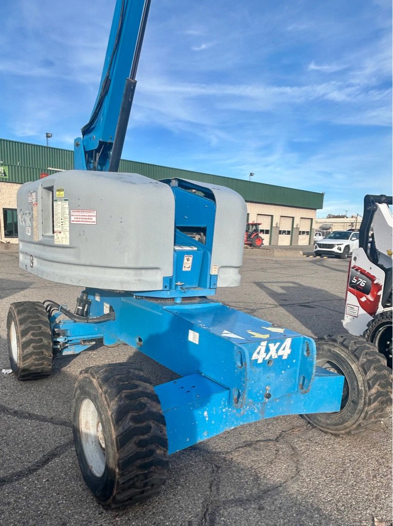 Blue 4x4 boom lift with large tires and extended arm, parked on asphalt in the equipment and trailer yard