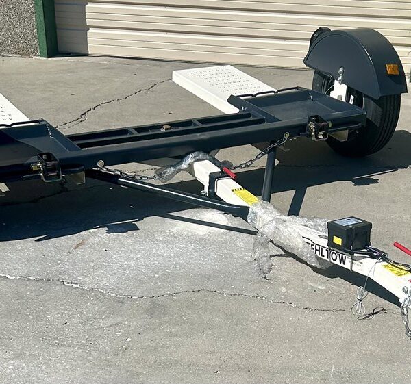 Close up of a black car tow dolly with white wheel platforms and fenders, sitting on a concrete lot in front of a garage building.