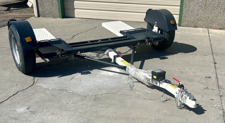 Close up of a black car tow dolly with white wheel platforms and fenders, sitting on a concrete lot in front of a garage building.