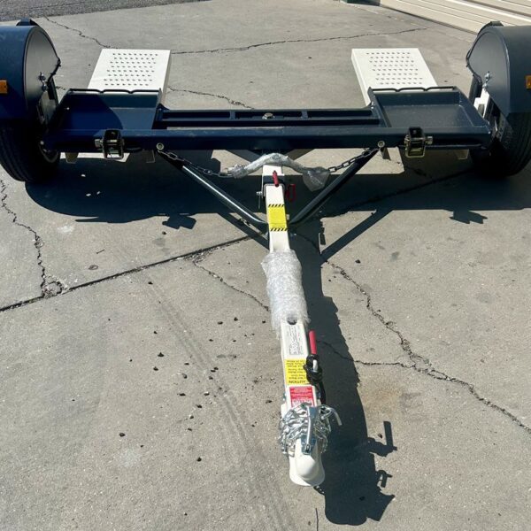 Car tow dolly with two ramps, black frame, and white hitch arm parked on gravel surface