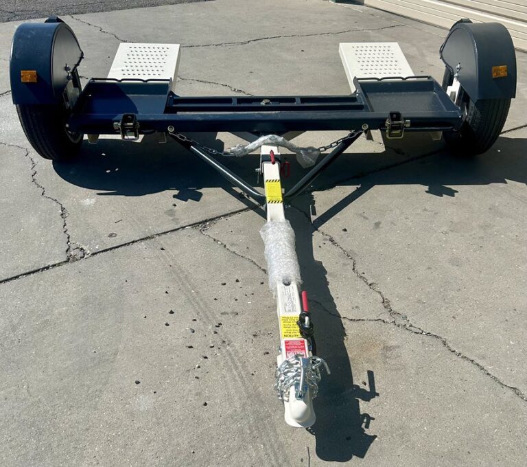Car tow dolly with two ramps, black frame, and white hitch arm parked on gravel surface