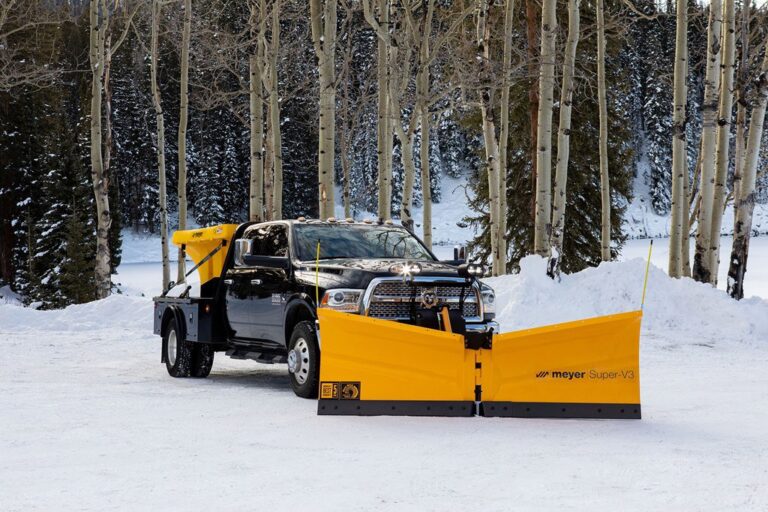 Black truck with yellow Meyer Super-V3 snow plow attachment parked on snowy ground among leafless trees with snow-covered forest background.
