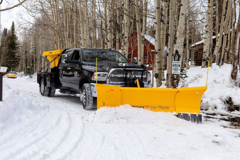 Black pickup truck equipped with a large yellow snow plow and a yellow salt spreader on the back, clearing snow on a snowy residential road