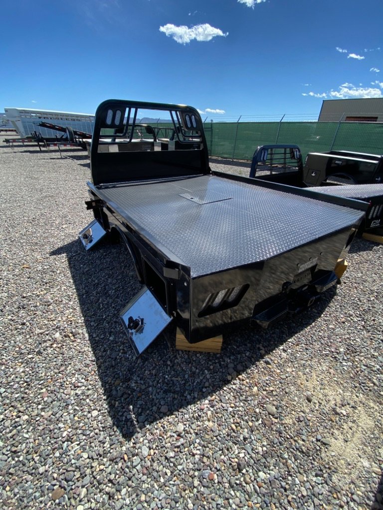 Black steel flatbed truck bed with diamond plate deck and headache rack in a mostly empty gravel trailer lot