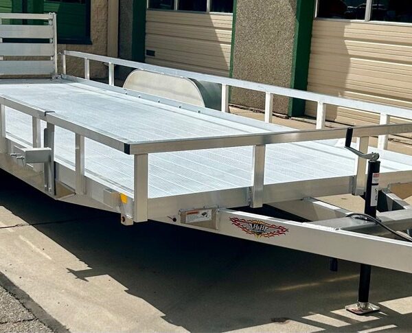 Large aluminum utility trailer with open sides, black wheel, and rear ramp gate, parked on concrete near a building.