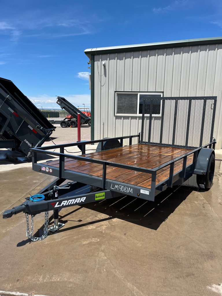 Freshly washed gray Lamar utility trailer with wood deck, mesh ramp gate, and single axle parked on wet concrete