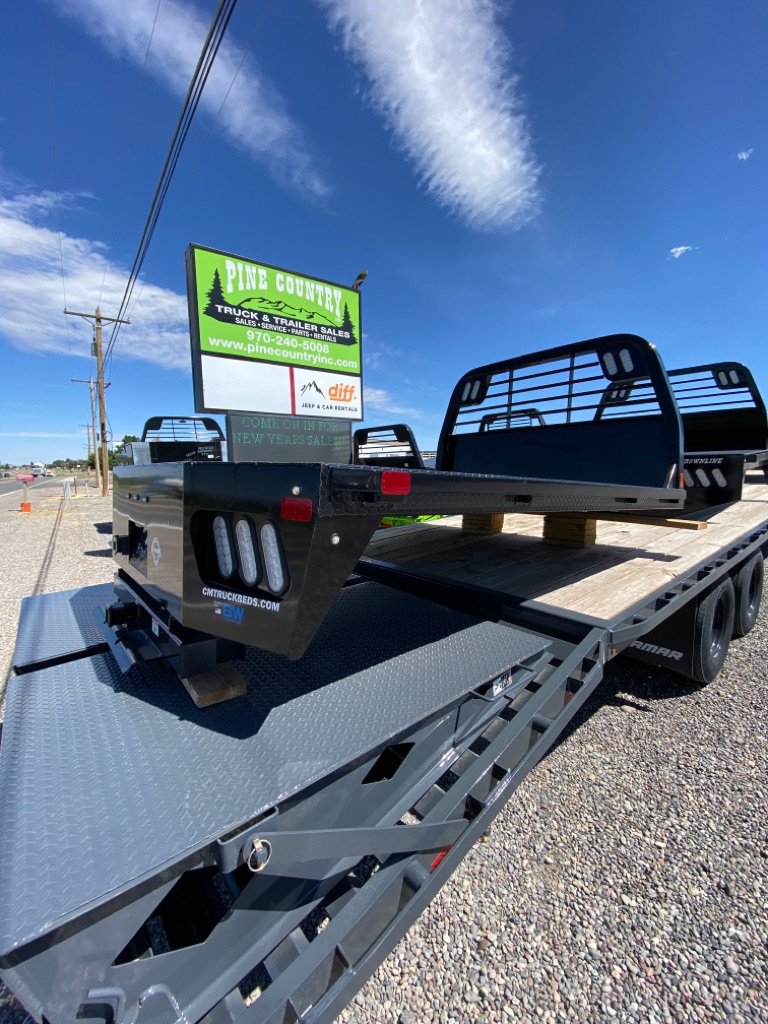Black truck beds stacked on flatbed trailer in gravel lot near Pine Country Trailer Sales sign