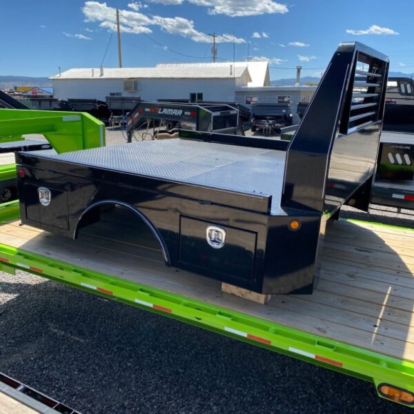 Black steel truck bed with diamond plate deck and toolboxes on green flatbed trailer in gravel lot