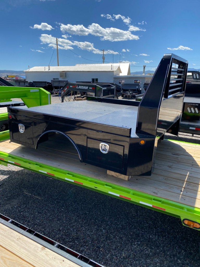 Black steel truck bed with diamond plate deck and toolboxes on green flatbed trailer in gravel lot