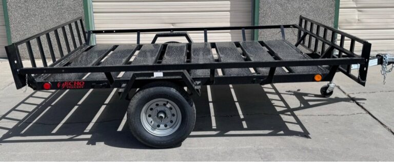 Black Echo utility trailer with metal grate floor and ramp gate, parked on concrete by beige garage doors.
