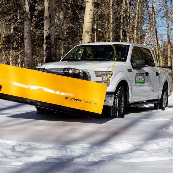 White pickup truck with yellow Meyer Drive Pro snow plow driving on snowy forest road lined with tall leafless trees.