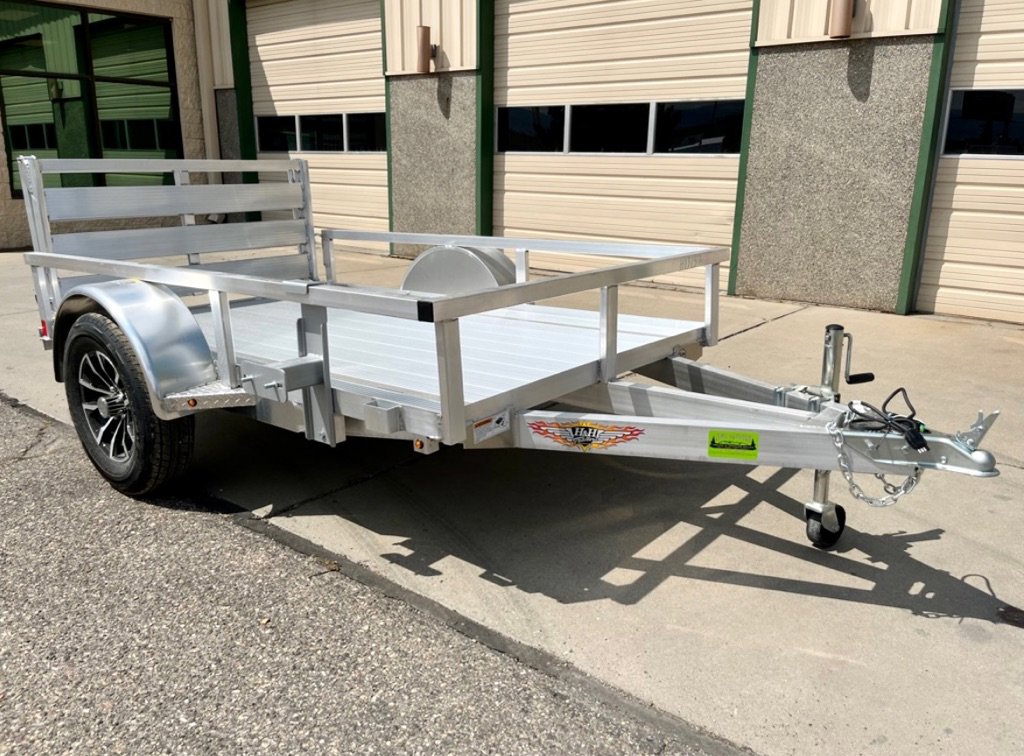 Aluminum utility trailer with open sides, black wheel, and rear ramp gate, parked on concrete near a building.