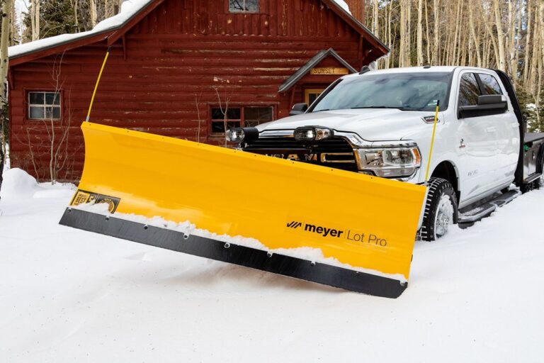 White truck with yellow Meyer Lot Pro snow plow clearing snow in front of red log cabin.