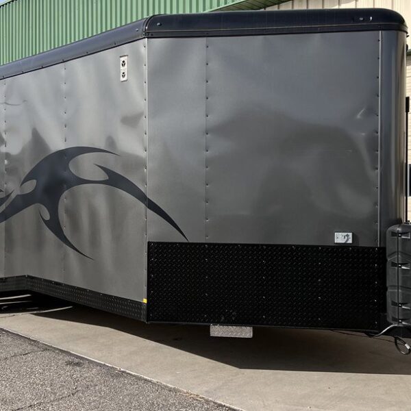 Large gray enclosed cargo trailer with black flame graphic on side, dual axles, black diamond plate trim, and black propane tanks, parked near building.