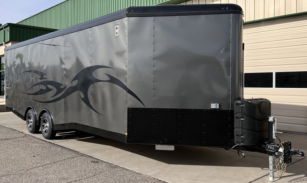 Large gray enclosed cargo trailer with black flame graphic on side, dual axles, black diamond plate trim, and black propane tanks, parked near building.