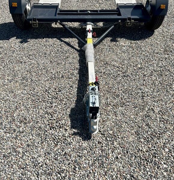 Black tow dolly with white ramps and a central hitch arm parked on gravel under bright sunlight.
