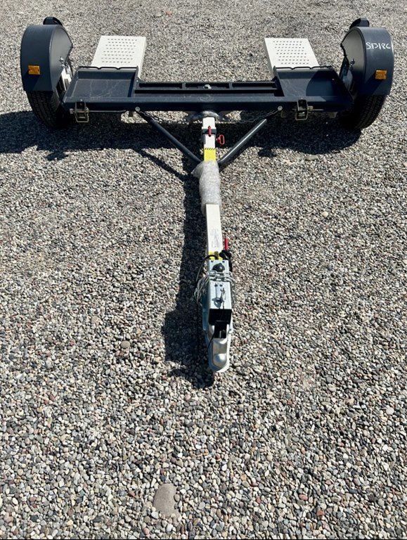 Black tow dolly with white ramps and a central hitch arm parked on gravel under bright sunlight.