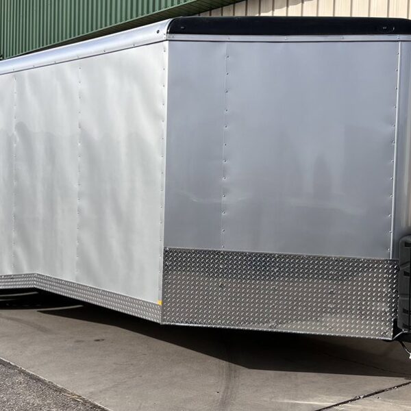 Long silver enclosed trailer with tandem axles and diamond plate trim parked outside garage doors