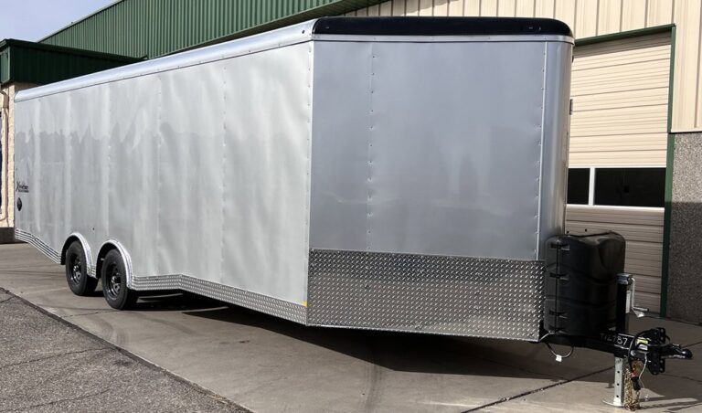 Long silver enclosed trailer with tandem axles and diamond plate trim parked outside garage doors