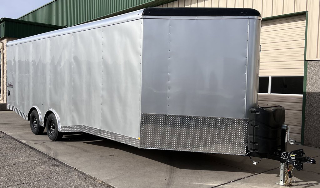Long silver enclosed trailer with tandem axles and diamond plate trim parked outside garage doors