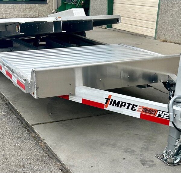 Aluminum Timpte car hauler trailer with tilt bed, red and white reflective tape, parked on concrete near garage doors.